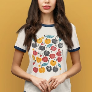 May include: White t-shirt with a blue ringer neckline featuring a colorful cherry print. The cherries are red, yellow, and black with green leaves.