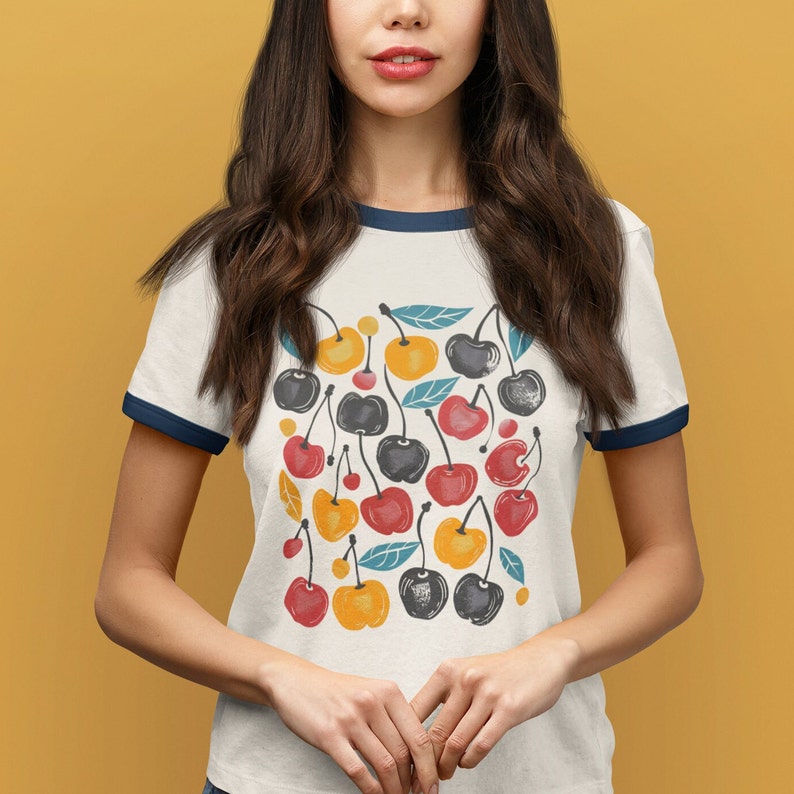 May include: White t-shirt with a blue ringer neckline featuring a colorful cherry print. The cherries are red, yellow, and black with green leaves.