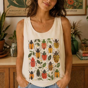 Puede incluir: Una camiseta sin mangas de color crema con un estampado gráfico central de varios escarabajos en tonos naranja, rojo, amarillo y verde, intercalados con pequeñas hojas verdes. La prenda tiene un cuello redondo amplio y un corte holgado.