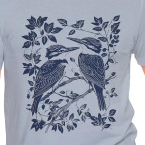 Op de afbeelding: Lichtblauw T-shirt met een donkerblauwe illustratie van twee vogels op een tak, omringd door bladeren. Het gedetailleerde ontwerp toont de vogels in een naturalistische stijl. Het T-shirt heeft een natuurthema.