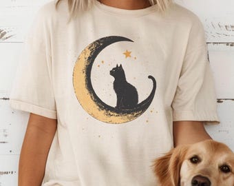 Op de afbeelding: Een crèmekleurig T-shirt met korte mouwen en een grafische print van een zwarte kat die op een getextureerde gele maansikkel zit. Het ontwerp bevat kleine gele sterretjes die rondom de maan en de kat zijn verspreid.