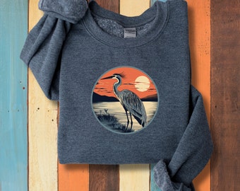 May include: A dark blue sweatshirt featuring a circular graphic of a heron standing in a lake at sunset. The heron is grey and white, and the sunset is orange and red.