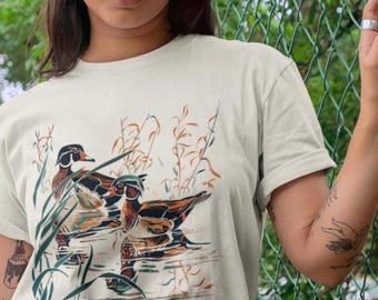Puede incluir: Una camiseta de manga corta de color crema con un estampado gráfico de dos patos joyuyo nadando entre juncos altos. La ilustración utiliza tonos tierra de marrón, naranja y verde para representar a los patos y la vegetación acuática.