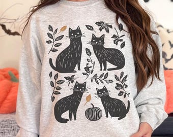 May include: A light gray sweatshirt featuring a black cat design with leaves and a pumpkin. The graphic includes four black cats, leaves, and a pumpkin, perfect for Halloween or cat lovers.