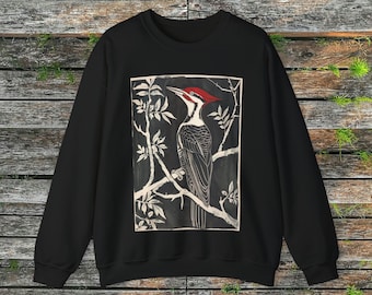 May include: Black sweatshirt featuring a graphic print of a woodpecker perched on a branch. The woodpecker is black and white with a red head and beak. The background is a dark grey.