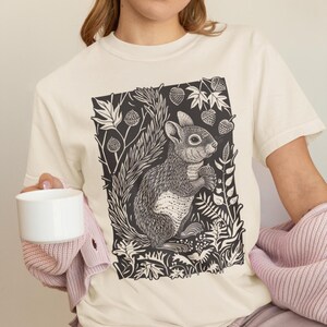 May include: A cream coloured t-shirt featuring a black and white illustration of a squirrel holding a nut in a woodland setting. The squirrel is surrounded by leaves, berries, and other woodland elements.