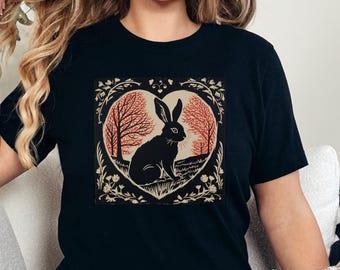 May include: A black short-sleeve t-shirt featuring a central graphic print of a black rabbit sitting inside a heart-shaped frame. The design includes orange-toned trees and foliage within the heart, surrounded by decorative floral line art.