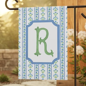 May include: A white garden flag with a blue and green floral pattern and a blue frame with the letter "R" in green. The flag is attached to a black metal pole.