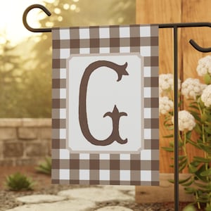 May include: A brown and white checkered garden flag with a large brown letter "G" in the center. The flag is attached to a black metal pole.