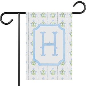 May include: A garden flag with a white background and a blue floral pattern. The flag features a large, light blue letter "H" inside a blue frame. The flag is hanging on a black metal stand.