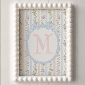 May include: Framed wall art with a white beaded frame. The artwork features a pink letter "M" within a blue ribbon design, set against a striped floral background. The background combines pink, blue, and floral patterns.