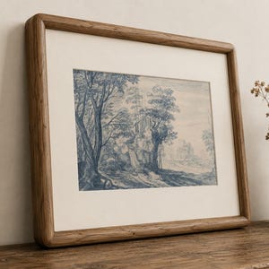 May include: A framed landscape print featuring a blue and white illustration of trees and a building. The artwork is set within a light-colored mat and a wooden frame. The frame is resting on a wooden surface.