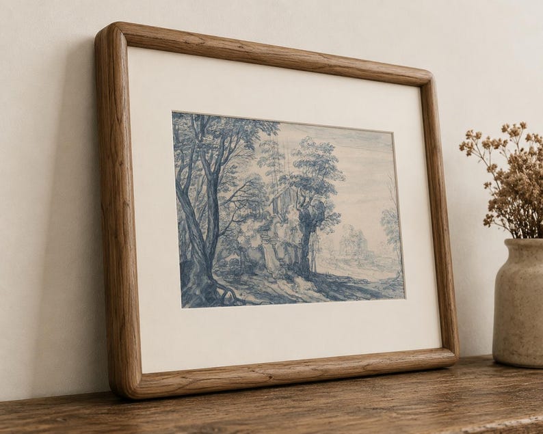 May include: A framed landscape print featuring a blue and white illustration of trees and a building. The artwork is set within a light-colored mat and a wooden frame. The frame is resting on a wooden surface.