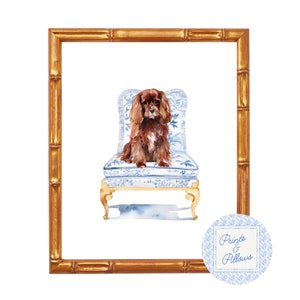 Könnte beinhalten: Aquarellgemälde eines braunen Cavalier King Charles Spaniels, der auf einem blau-weiß gemusterten Stuhl sitzt. Der Stuhl hat goldene Beine und goldene Verzierungen. Das Gemälde ist in einem goldenen Bambusrahmen gerahmt. Das Bild enthält den Text "Prints & Pillows".