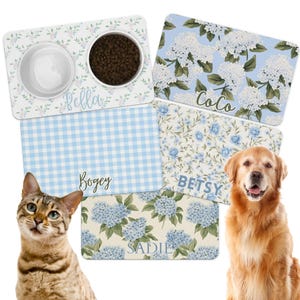 May include: A collection of personalized pet food mats and bowls. The mats feature floral, gingham, and hydrangea patterns in blue and white. Names like "Bella", "Coco", "Bogey", "Betsy", and "Sadie" are displayed. A cat and a golden retriever are also in the image.