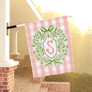 May include: A pink and white gingham garden flag with a decorative floral crest. The crest features a pink letter "S" surrounded by green leaves and a green bow. The flag is hanging outside.