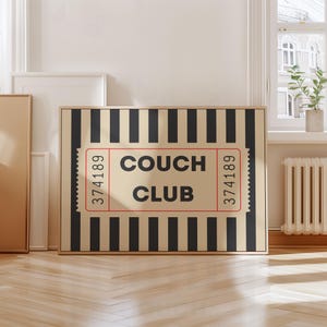 Op de afbeelding: Een ingelijste kunstdruk met een beige en zwarte gestreepte achtergrond. De print heeft een ticket-achtig ontwerp met de woorden "COUCH CLUB" in vette letters. Het ticket heeft aan weerszijden het nummer "374189".