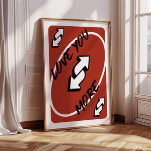Può includere: Stampa artistica incorniciata con un design di carta rossa, con la scritta "LOVE YOU MORE" e frecce bianche. La stampa è in una cornice dorata, appoggiata a un muro bianco e un pavimento in legno.
