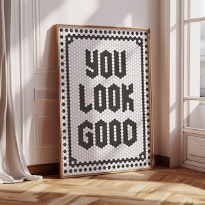 May include: A framed art print with the words "YOU LOOK GOOD" in black block letters. The letters are made of small, black and white hexagonal tiles. The frame is a light brown color, and the print is leaning against a white wall.