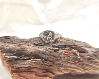 May include: A silver signet ring with a rectangular face featuring an engraved design of a bird and floral elements. The ring is displayed on a piece of weathered wood, showcasing its details.