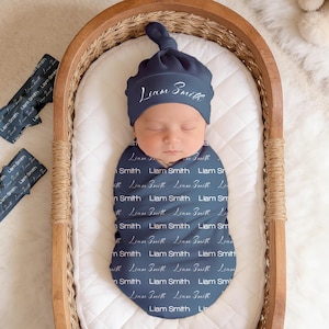 May include: A baby wrapped in a navy blue swaddle and hat, both featuring the name "Liam Smith" in white script. The baby rests in a wooden cradle with a white, quilted lining. Two matching headbands are also present. The cradle is made of wood.