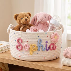 以下が含まれることがあります： クリーム色のロープバスケットに、カラフルな文字で「Sophia」と書かれています。バスケットには、テディベア、ピンクのウサギ、おしゃぶり、哺乳瓶が入っています。ピンクの花が側面を飾っています。