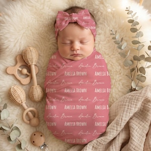 May include: A baby wrapped in a pink swaddle blanket and matching headband, both printed with the name "Amelia Brown" in a repeating pattern. The baby is lying on a fluffy white surface, surrounded by wooden toys and eucalyptus sprigs.