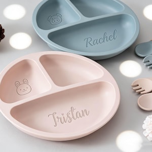 Op de afbeelding: Twee verdeelde babyborden, één lichtroze met een konijn en de naam 'Tristan', de andere blauw met een varken en de naam 'Rachel'. Inclusief bijpassend bestek en een leeuwenrammelaar.