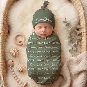 May include: A baby wrapped in a green swaddle and hat, both featuring the name "Emma Jones" in white script. The baby lies in a woven basket, with wooden toys. The blanket and hat are olive green.