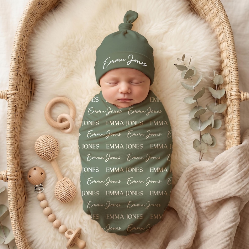 K&ouml;nnte beinhalten: Ein salbeigr&uuml;nes Baby-Pucktuch und eine passende Knotenm&uuml;tze, beide mit dem Namen Emma Jones in einem sich wiederholenden Muster aus wei&szlig;er Schreib- und Serifenschrift bedruckt, pr&auml;sentiert mit einer Holzrassel und einem Schnullerhalter.