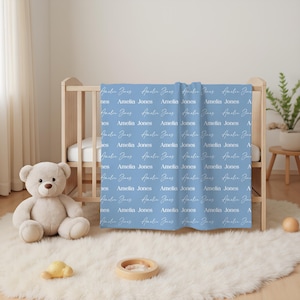 K&ouml;nnte beinhalten: Eine hellblaue Babydecke mit einem sich wiederholenden Muster des Namens Amelia Jones, gedruckt in abwechselnden wei&szlig;en Serif- und Schreibschriften. Die Decke h&auml;ngt &uuml;ber der Seite eines Holzkinderbetts, in der N&auml;he eines Pl&uuml;schb&auml;ren.