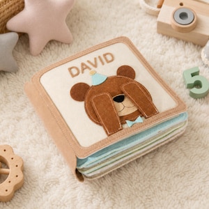 May include: A square fabric book with a cream-colored cover featuring a brown felt bear wearing a blue party hat and bow tie. The name DAVID is stitched in brown letters above the bear. The book has light blue and cream pages visible on the side.
