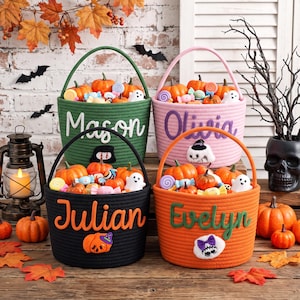 May include: Four round woven rope baskets for Halloween, each featuring a handle and a personalized name in cursive script. The baskets are green, pink, black, and orange, filled with small pumpkins and candies, and decorated with felt appliques.