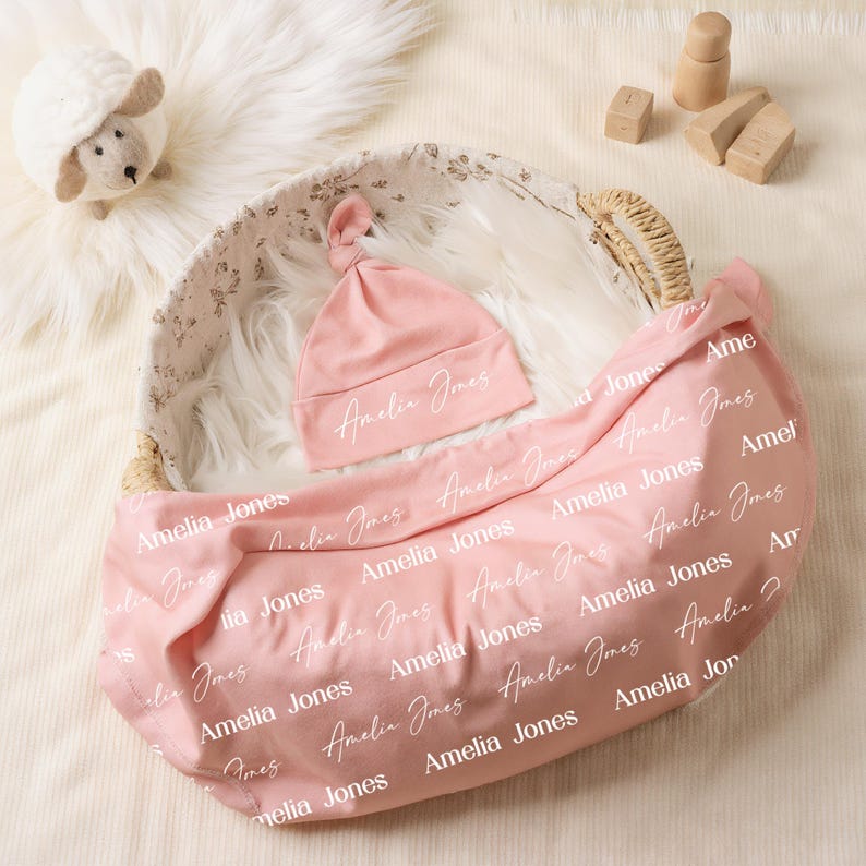 K&ouml;nnte beinhalten: Eine weiche rosa Babym&uuml;tze mit Knoten und eine passende rosa Wickeldecke, beide mit dem Namen Amelia Jones in einem sich wiederholenden wei&szlig;en Schrift- und Serifenschriftmuster bedruckt, in einem geflochtenen Korb mit floralem Stofffutter.