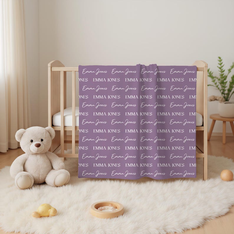 K&ouml;nnte beinhalten: Eine violette Decke mit einem sich wiederholenden Muster des Namens Emma Jones, gedruckt in abwechselnden Schreib- und Serifenschriften. Die Decke liegt &uuml;ber einem Holzbettchen, mit einem Teddyb&auml;ren auf einem wei&szlig;en Teppich im Vordergrund.