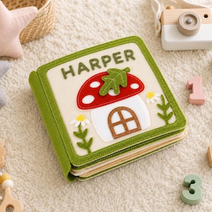 May include: A square fabric quiet book with a cream-colored cover featuring the name HARPER in green letters. The cover displays a red and white mushroom house with a green leaf on top, flanked by two small white daisies with green stems.