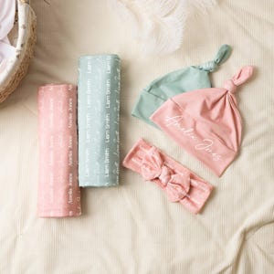 K&ouml;nnte beinhalten: Zwei gefaltete Wickeldecken, eine rosa mit dem Namen Amelia Jones und eine salbeigr&uuml;ne mit dem Namen Liam Smith. Zwei geknotete Babym&uuml;tzen in passendem Rosa und Salbeigr&uuml;n, mit Amelia Jones auf der rosa M&uuml;tze und ein rosa Schleifen-Stirnband.