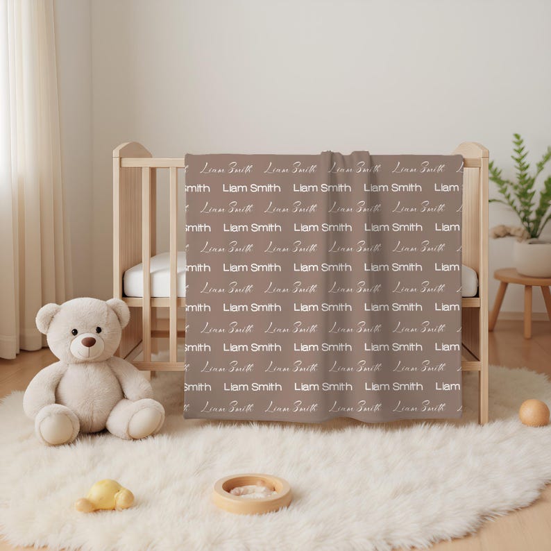 K&ouml;nnte beinhalten: Eine taupefarbene Babydecke mit einem sich wiederholenden Muster des Namens Liam Smith, der in einer wei&szlig;en, stilisierten Schreibschrift gedruckt ist. Die Decke h&auml;ngt &uuml;ber der Seite eines Holzkinderbetts neben einem hellbraunen Pl&uuml;schb&auml;ren.