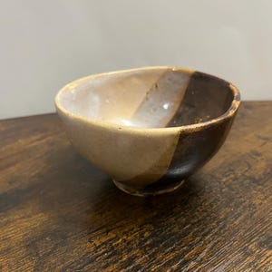 Puede incluir: Un pequeño cuenco de cerámica con un diseño bicolor que presenta un acabado liso de color beige claro en un lado y un esmalte marrón oscuro brillante en el otro. El cuenco tiene un borde irregular y orgánico, además de una base circular pequeña.