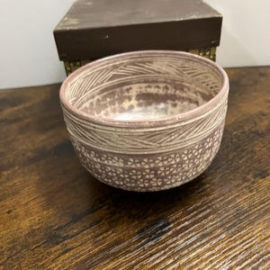 May include: A round ceramic bowl with a mauve and cream color palette. The exterior features a band of etched diagonal lines near the rim and a repeating pattern of small, stamped flower shapes across the lower body. A brown box sits behind it.