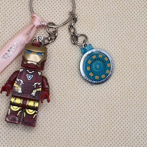 May include: Iron Man themed keychains. One keychain features an Iron Man figure in red, gold, and maroon. The other keychain is a blue and silver circular design with the text "That Tony Stark Has a Heart". A pink ribbon with text is also present.