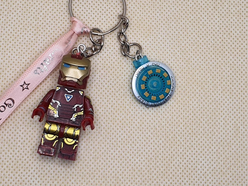 May include: Iron Man themed keychains. One keychain features an Iron Man figure in red, gold, and maroon. The other keychain is a blue and silver circular design with the text "That Tony Stark Has a Heart". A pink ribbon with text is also present.