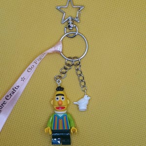 Op de afbeelding: Een sleutelhanger met een Bert-figuur en een witte vogelbedel. De Bert-figuur heeft een geel gezicht, zwart haar en een kleurrijk gestreept shirt. De sleutelhanger heeft een stervormige sluiting en een roze lint met de tekst "Sure Crafts Go Figur".