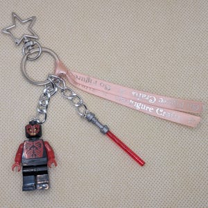 Puede incluir: Un llavero con una figura de Lego de Darth Maul, con un sable de luz rojo y una cinta rosa que dice "Figure Crafts". El llavero tiene un cierre plateado en forma de estrella y una cadena.
