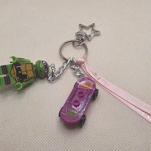 May include: A keyring featuring a green turtle character figurine, a purple plastic skateboard charm, and a light pink ribbon with the text Go Figure Crafts printed in white. The charms are attached to a silver metal ring with a star-shaped clasp.