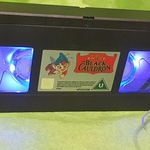 May include: A black VHS tape with the title "The Black Cauldron" and a cartoon character on the label. The tape has blue lights illuminating the interior reels. The tape is on a green surface.