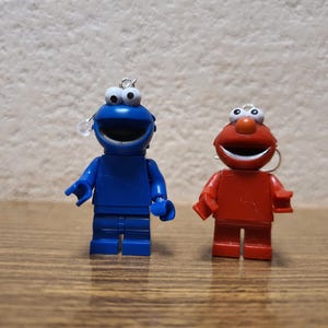 Könnte beinhalten: Zwei kleine Plastikfiguren der Sesamstraße-Charaktere Cookie Monster und Elmo. Cookie Monster ist blau mit großen weißen Augen und einem weit geöffneten Mund. Elmo ist rot mit einem großen Lächeln und weißen Augen. Beide Figuren haben eine kleine Schlaufe oben auf dem Kopf.