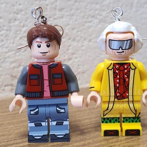 Pode incluir: Um par de brincos com minifiguras Lego de Doc Brown e Marty McFly da franquia de filmes De Volta para o Futuro. Doc Brown está vestindo uma jaqueta amarela, uma bandana vermelha e óculos de sol. Marty McFly está vestindo um colete vermelho, calças cinzas e uma camisa rosa.