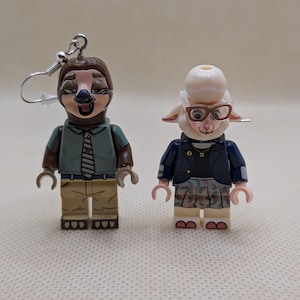 Puede incluir: Un par de pendientes hechos a mano con figuras estilo Lego. Un pendiente representa un personaje de perezoso con camisa verde y corbata. El otro pendiente muestra un personaje de oveja con gafas y chaqueta. Los pendientes están sujetos a ganchos plateados.