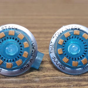 Puede incluir: Un par de pendientes redondos de plata y azul con un centro azul y un borde plateado. Los pendientes tienen el texto "PROOF THAT TONY STARK HAS A HEART" escrito alrededor del borde.